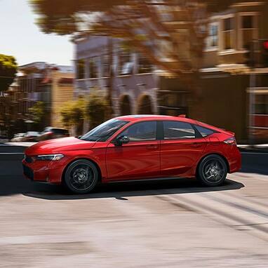 2027 Honda Civic Hatchback Sport Hybrid in Rallye Red profile driving on a city street (Compact Hatchback).